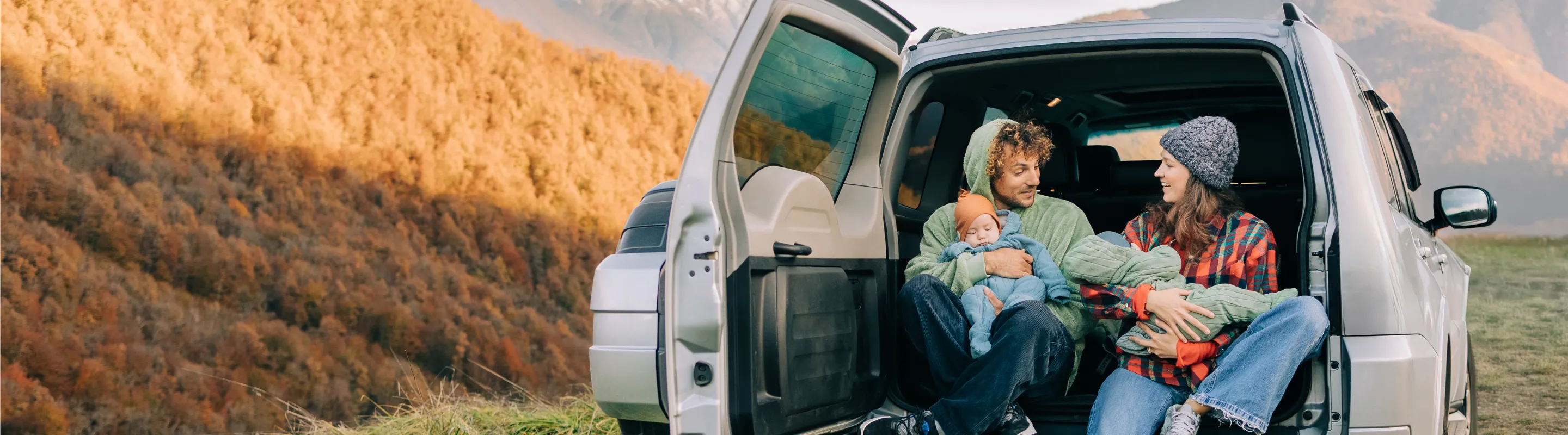 a mother and father with road trip travel insurance sit on the back of their SUV holding babies in the mountains  