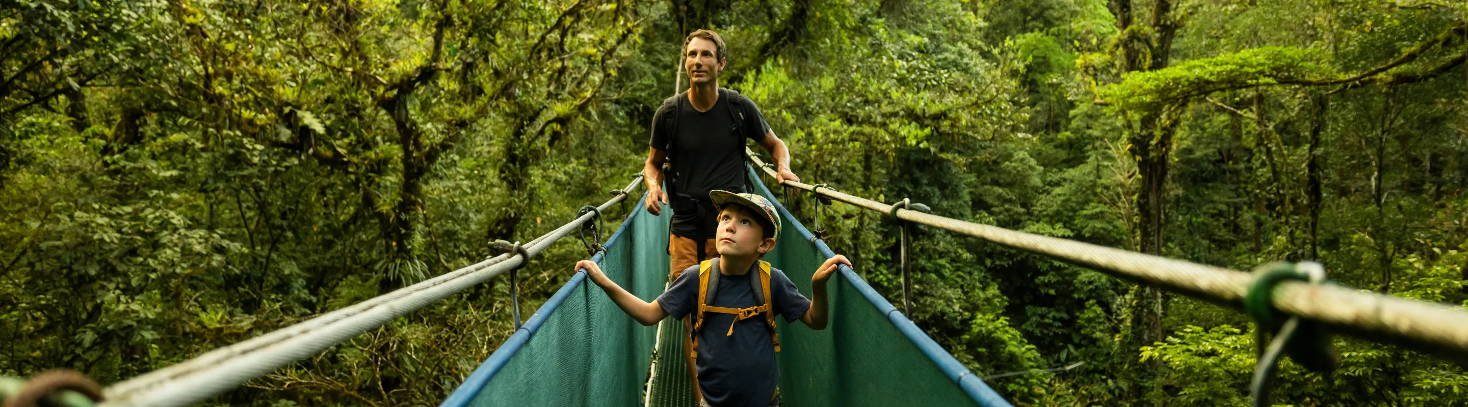 a father and son cross a suspension bridge in a tropical rainforest, insured with travel insurance for Costa Rica 