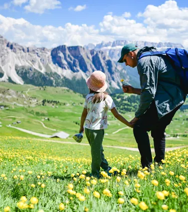 a father with travel insurance for Italy shows his daughter a flower as they hike the Italian Dolomites 