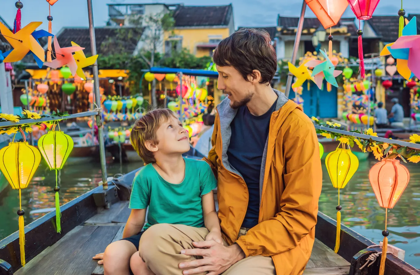 a father and son with travel insurance for Vietnam ride a traditional boat through Hoi An surrounded by colorful lanterns