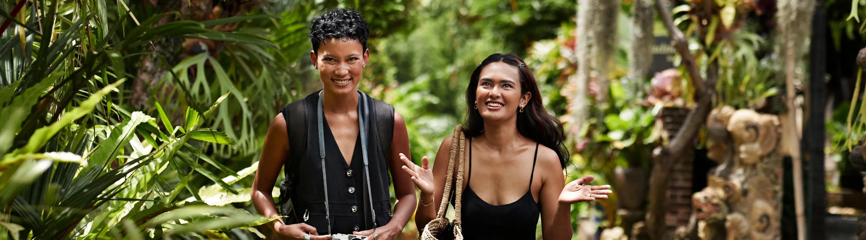 two female friends smile as they walk through a tropical garden, insured with travel insurance for Bali