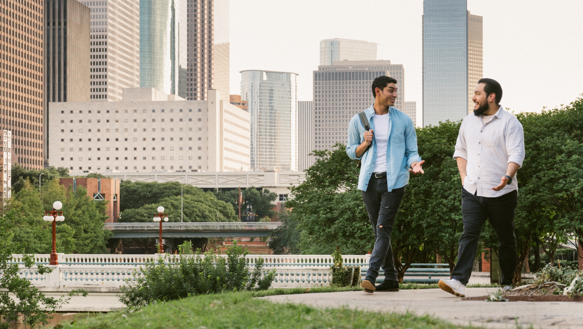 two male friends with travel insurance for hurricanes walk through a park near the Houston skyline 