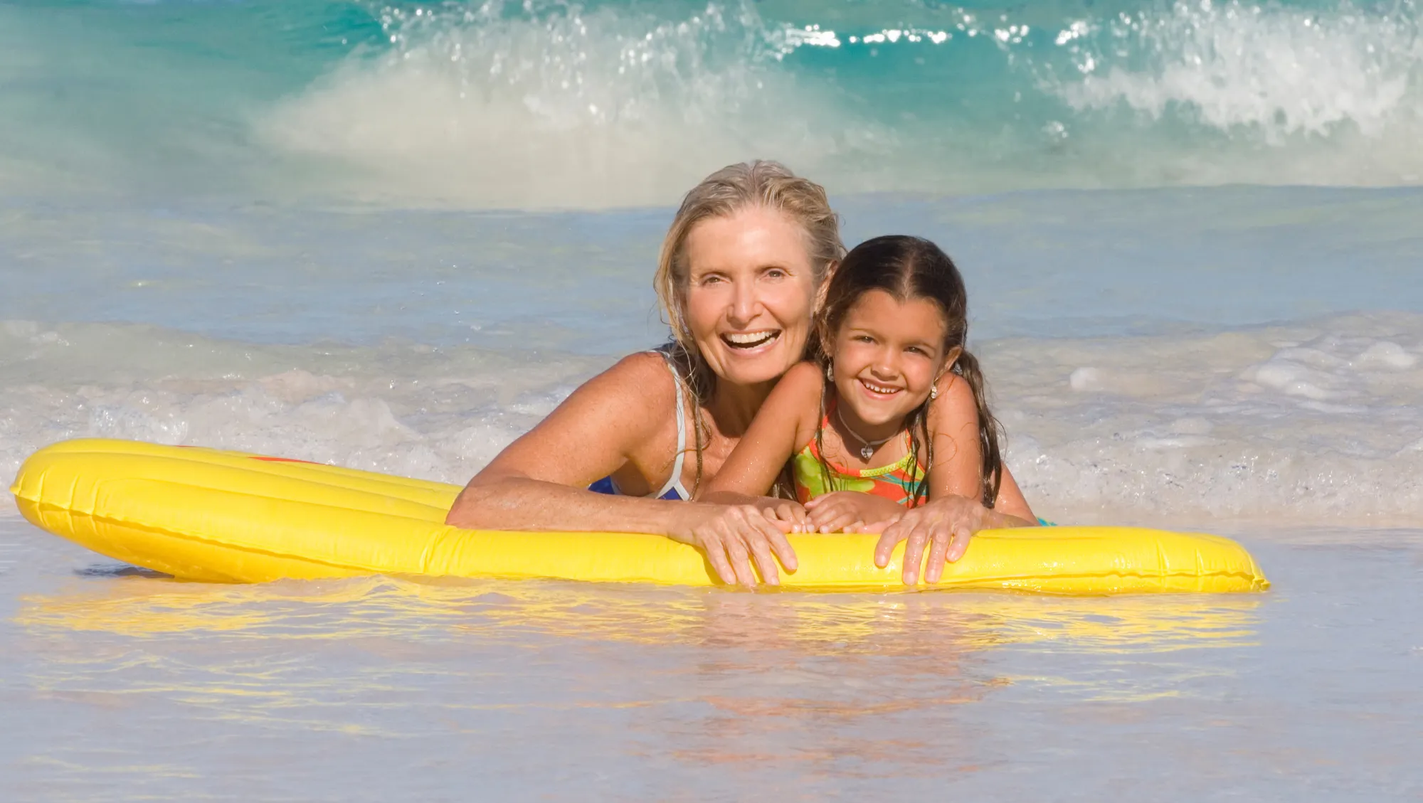 an adult and child with travel insurance for hurricanes lie on a yellow inflatable raft in shallow ocean water 