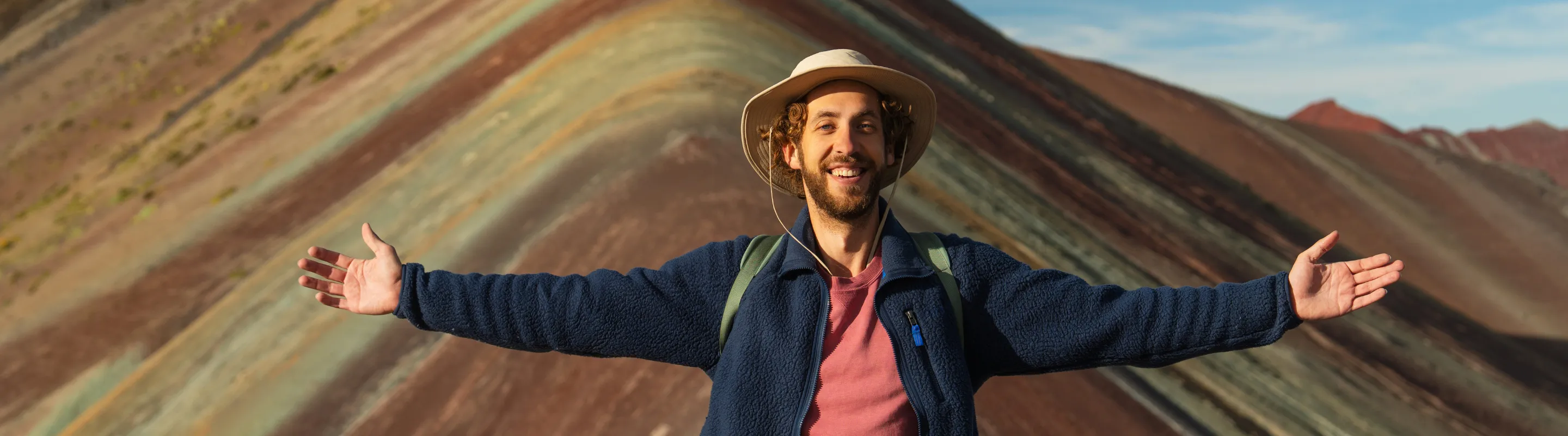 a young man stands and raises his arms at Rainbow Mountain, Peru, insured with travel insurance for South America 