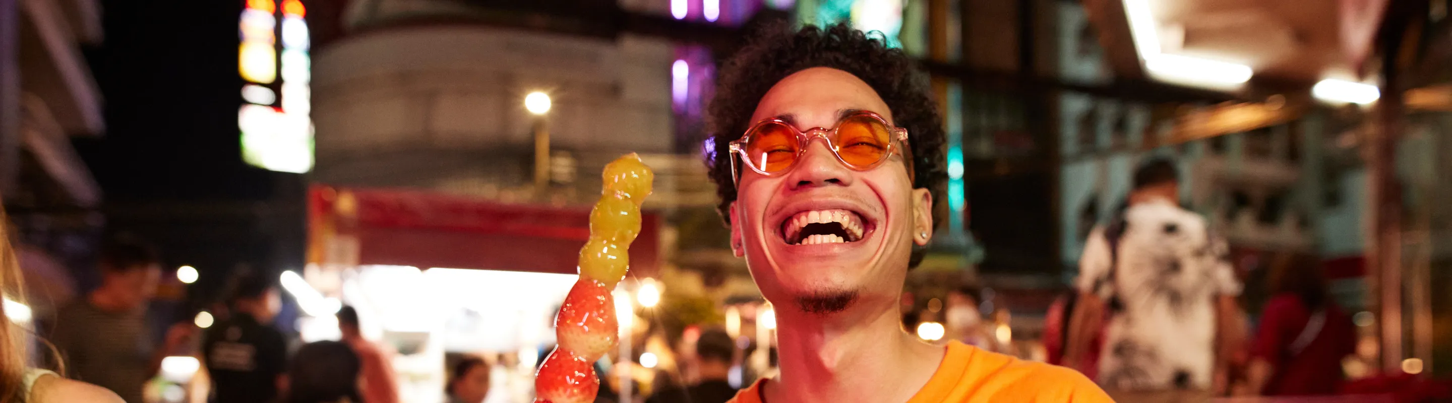 a man with travel insurance for culinary trips sits at an outdoor night market table holding a skewer of colorful candied fruit