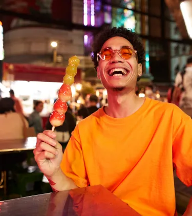 a man with travel insurance for culinary trips sits at an outdoor night market table holding a skewer of colorful candied fruit