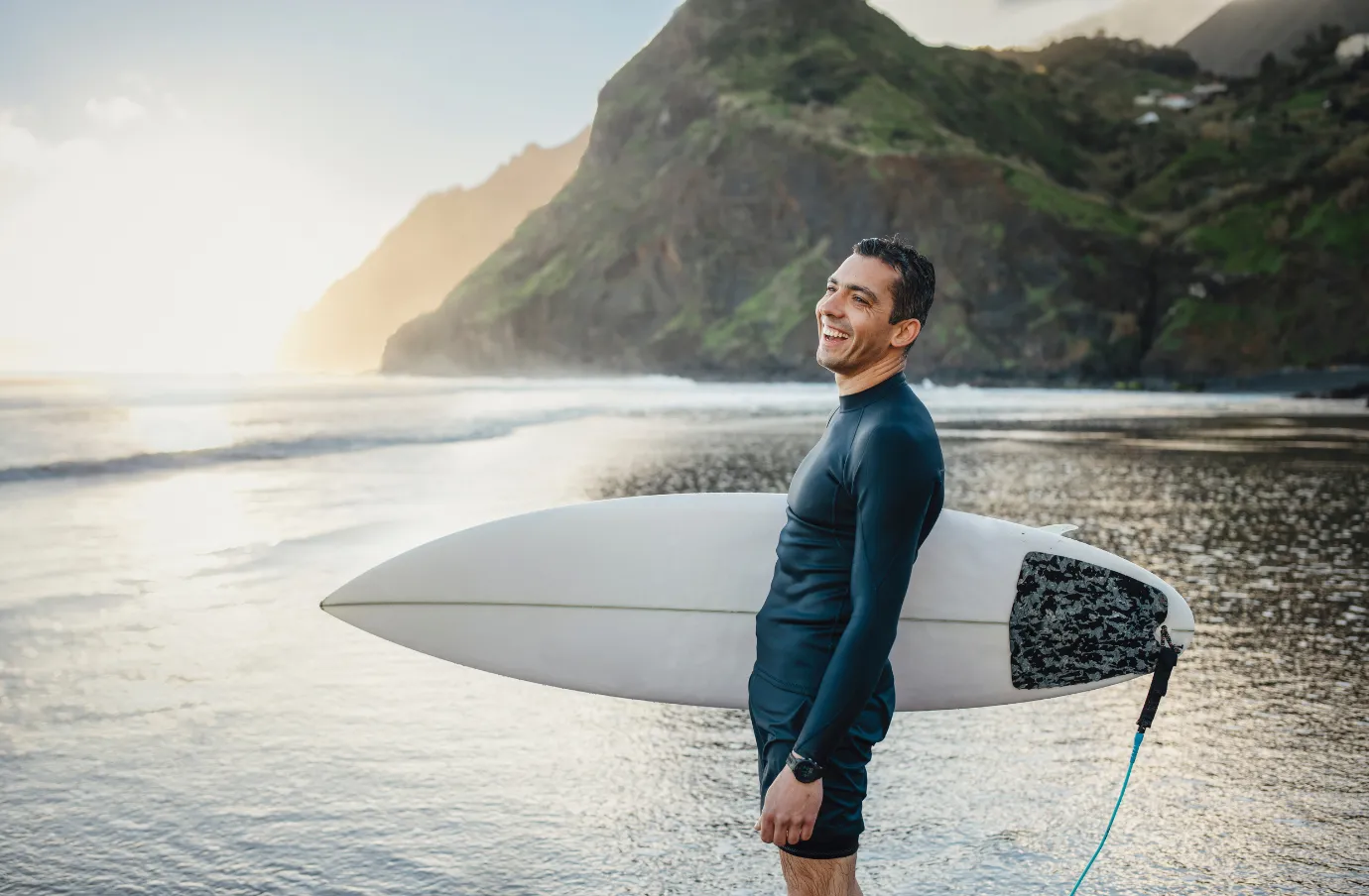 a middle-aged man holds a surfboard on a beach in Madeira, insured with travel insurance for Portugal