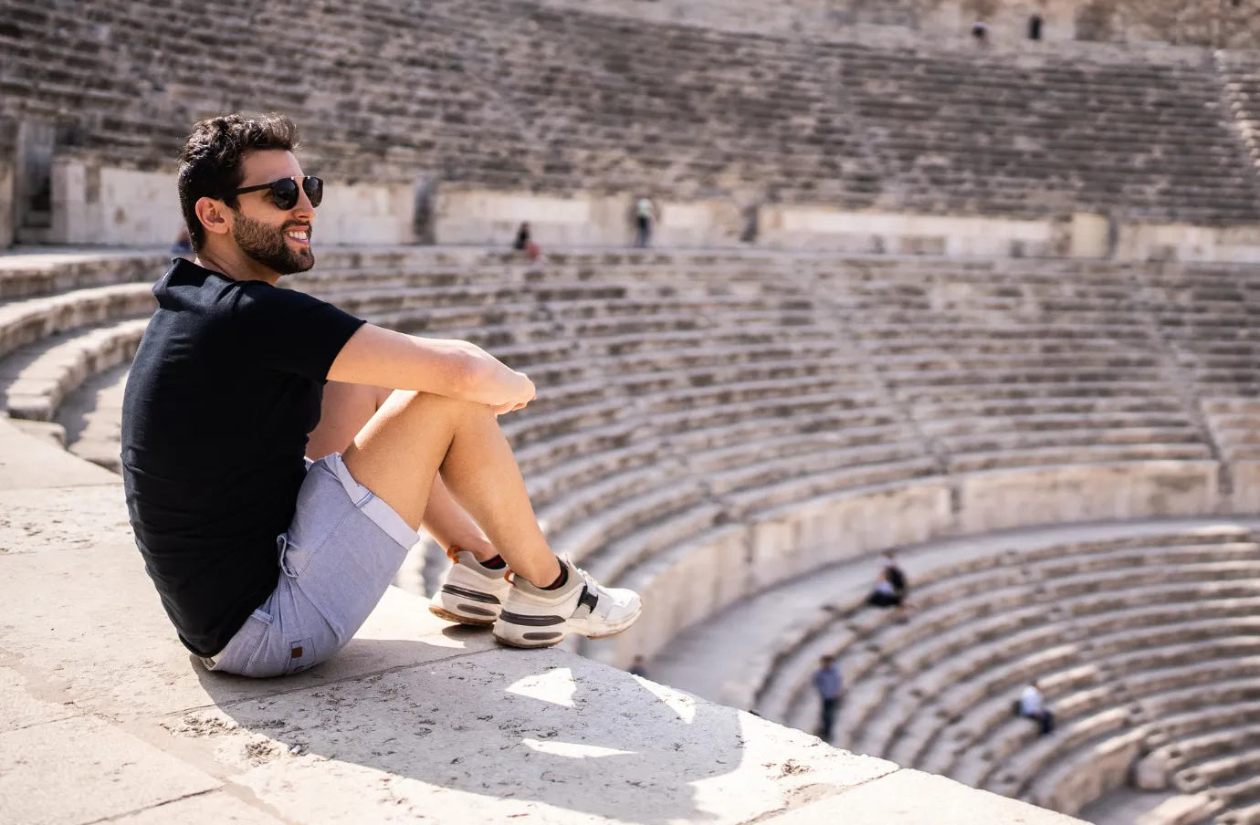 a young man with sunglasses and travel insurance for Jordan sits in an ancient amphitheater in Amman, Jordan