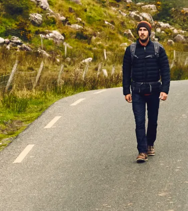 a young man with Ireland travel insurance walks down a scenic road near greenery and mountains 