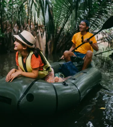a man and woman with international travel insurance share a kayak and paddle through a mangrove with palm fronds