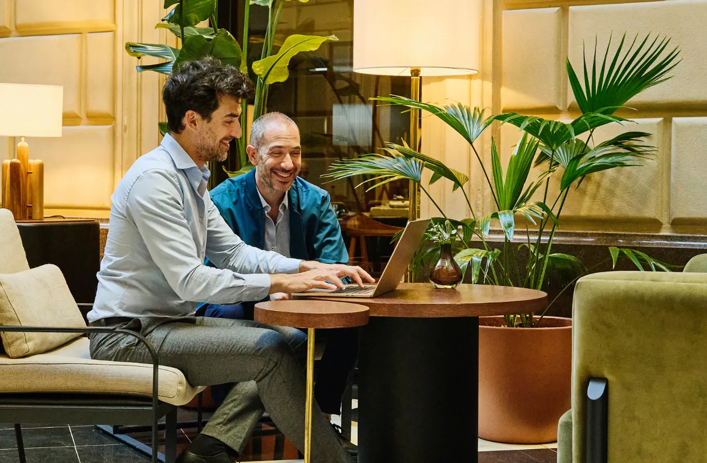 two men sit at a table looking at our travel insurance FAQs on a laptop in an indoor lounge