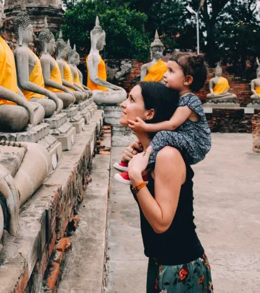 a mother with travel insurance for Thailand carries her young daughter while viewing rows of Buddha statues
