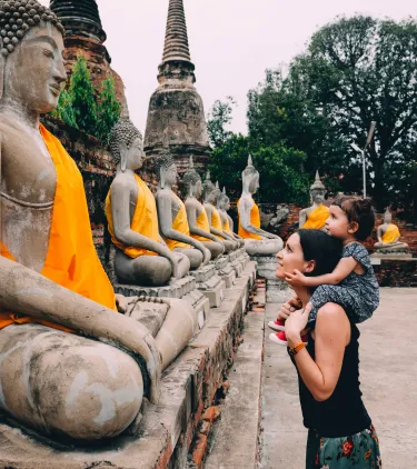 a mother with travel insurance for Thailand carries her young daughter while viewing rows of Buddha statues