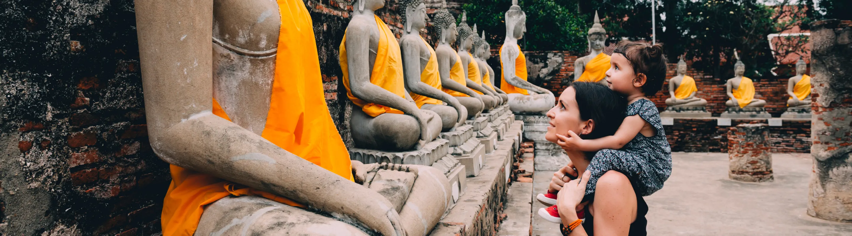 a mother with travel insurance for Thailand carries her young daughter while viewing rows of Buddha statues