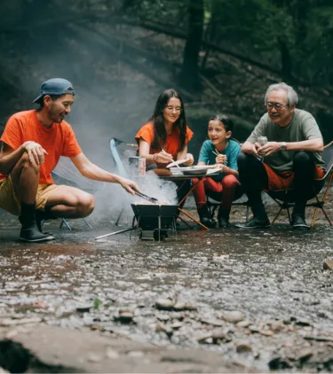 a multi-generational family with travel insurance for nature trips sits and cooks by the fire in the shallow water of a creek