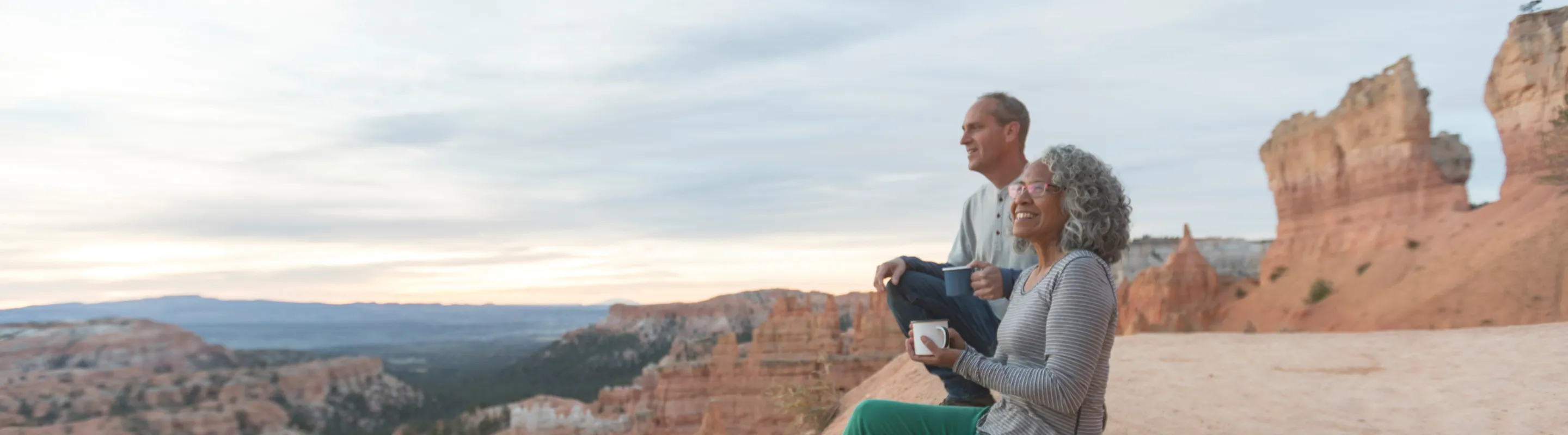 an older couple with domestic travel insurance sits on a red rock canyon's ledge in Utah and soaks up the lookout's views
