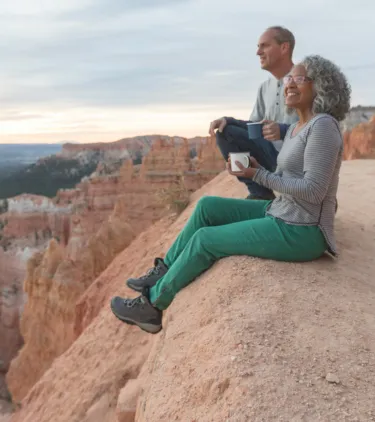 an older couple with domestic travel insurance sits on a red rock canyon's ledge in Utah and soaks up the lookout's views