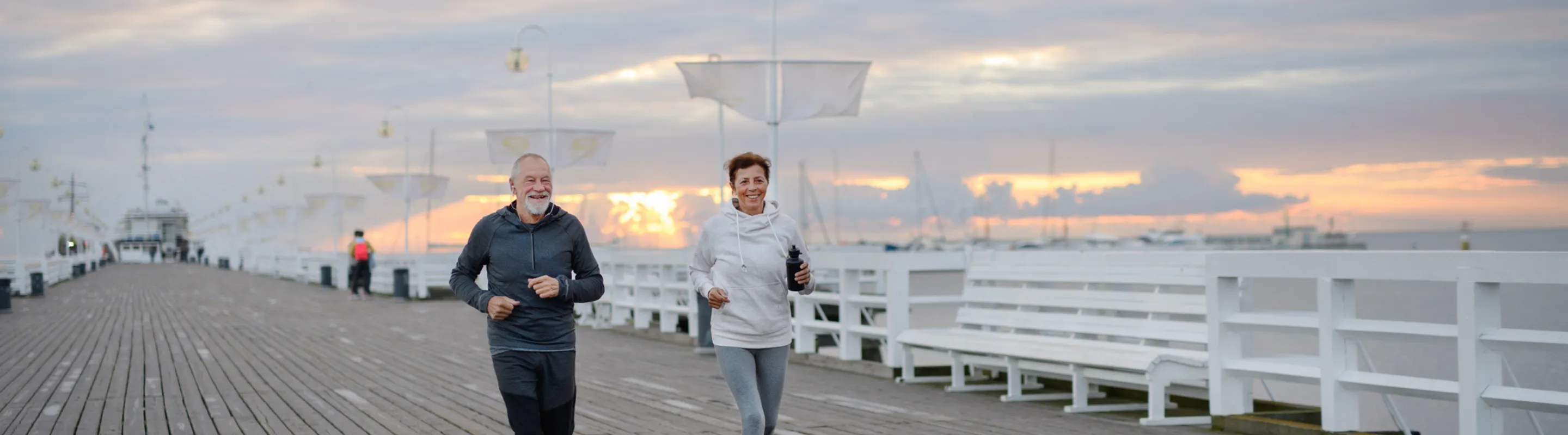 an older couple with travel insurance for missed connections jogs on a pier over the ocean at sunrise