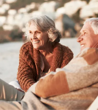 two older adults smile and sit in beach chairs along a shoreline on vacation after comparing travel insurance plans