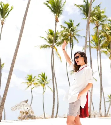 a woman with travel insurance for the Dominican Republic strolls through a palm-lined beach with huts in the background