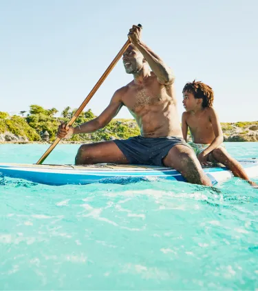 A father with travel insurance for the Caribbean paddleboards with a child seated behind in crystal-clear ocean water