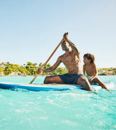 A father with travel insurance for the Caribbean paddleboards with a child seated behind in crystal-clear ocean water