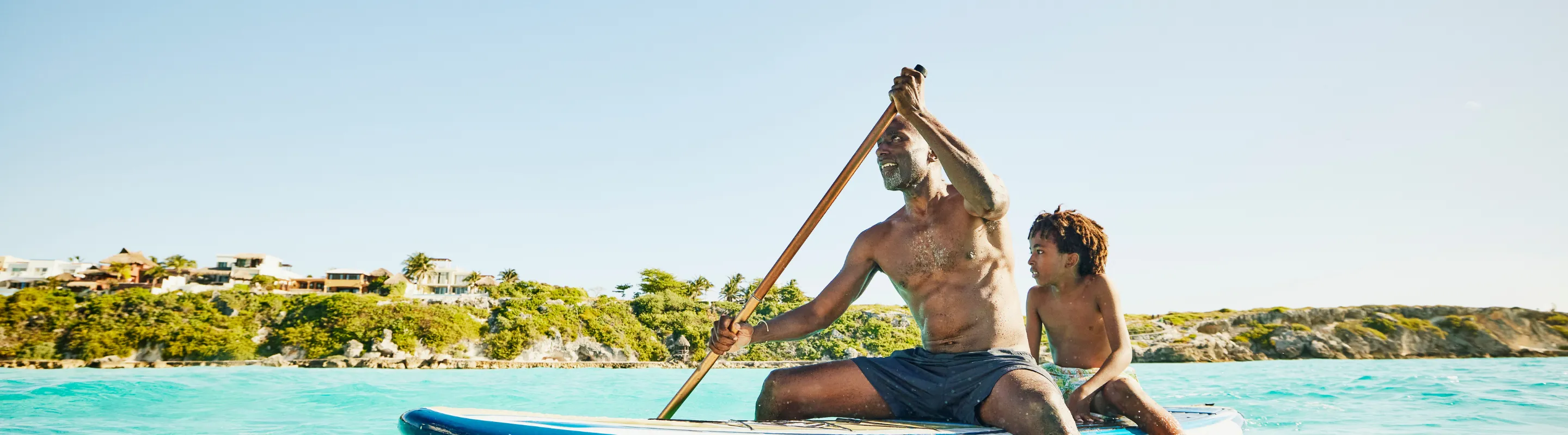 A father with travel insurance for the Caribbean paddleboards with a child seated behind in crystal-clear ocean water