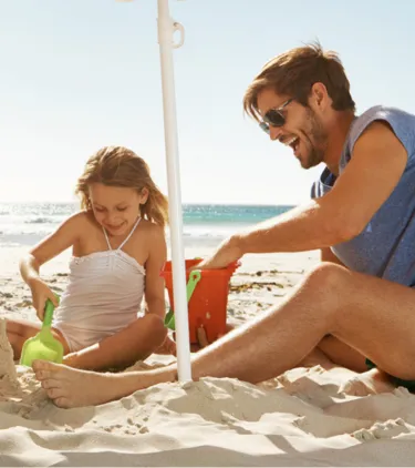 parents with travel insurance for beach trips help their young daughter build a sandcastle by the ocean on a sunny day
