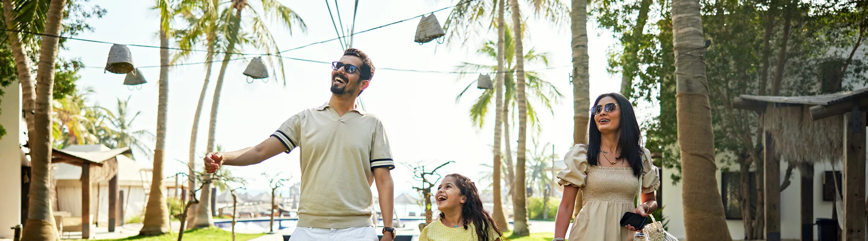 two young parents with long-term travel insurance walk alongside their daughter through a tropical resort with palm trees
