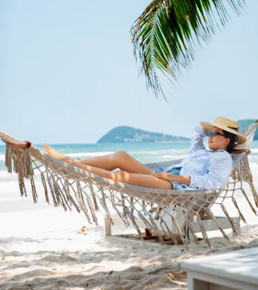 a woman with travel insurance for the Caribbean lounges in a hammock near the water's edge on a sandy beach