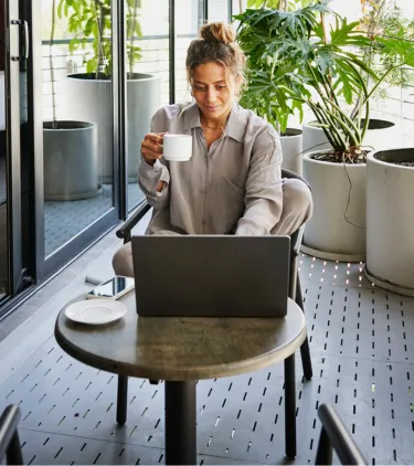 a remote worker with business travel insurance sits on a hotel balcony with a coffee and a laptop