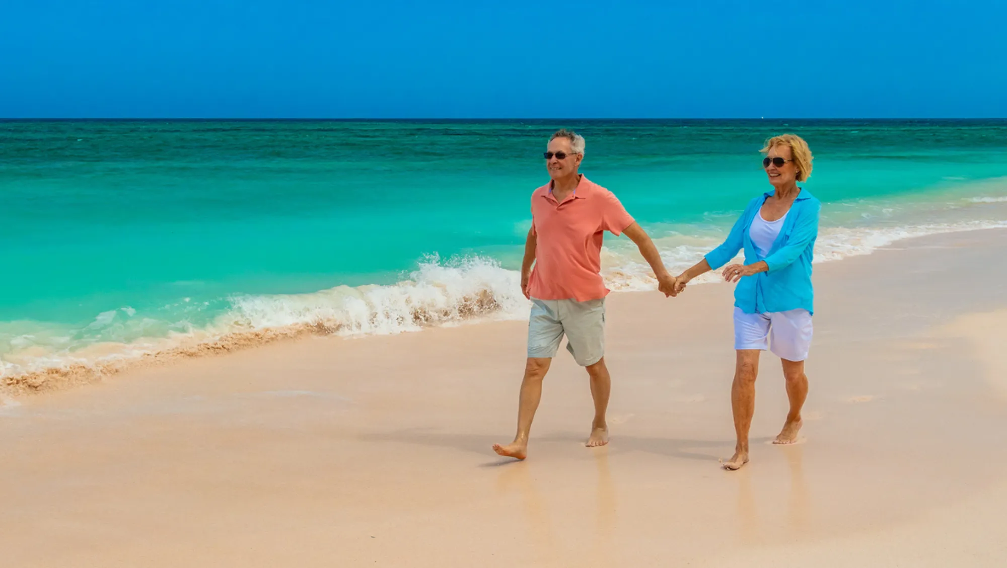 a senior couple holds hands while walking on a beach with turquoise water after asking, "Is travel insurance worth it?"