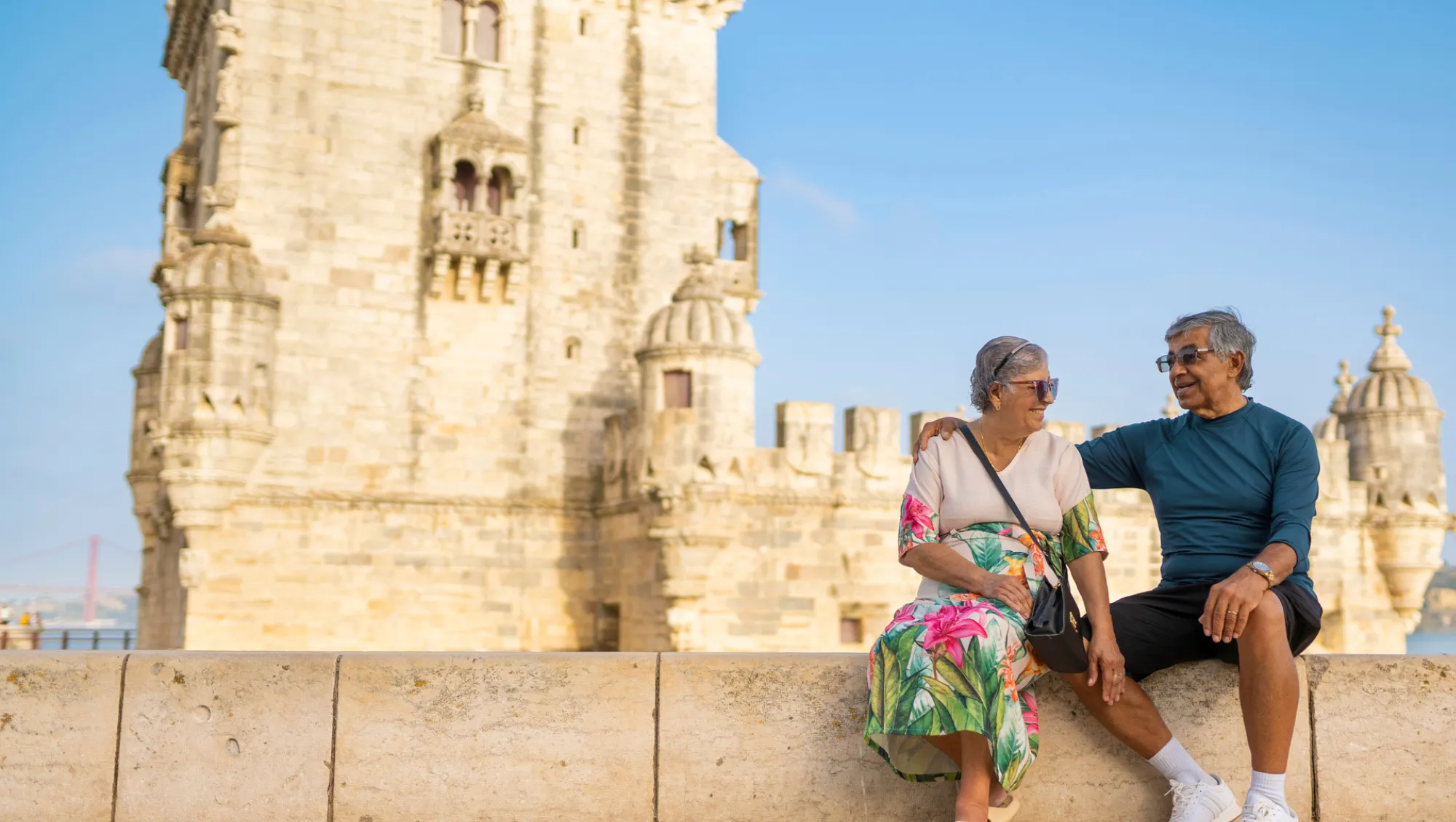 a senior couple sits in front of Belem Tower, Portugal, on vacation after asking, "Is travel insurance worth it?"