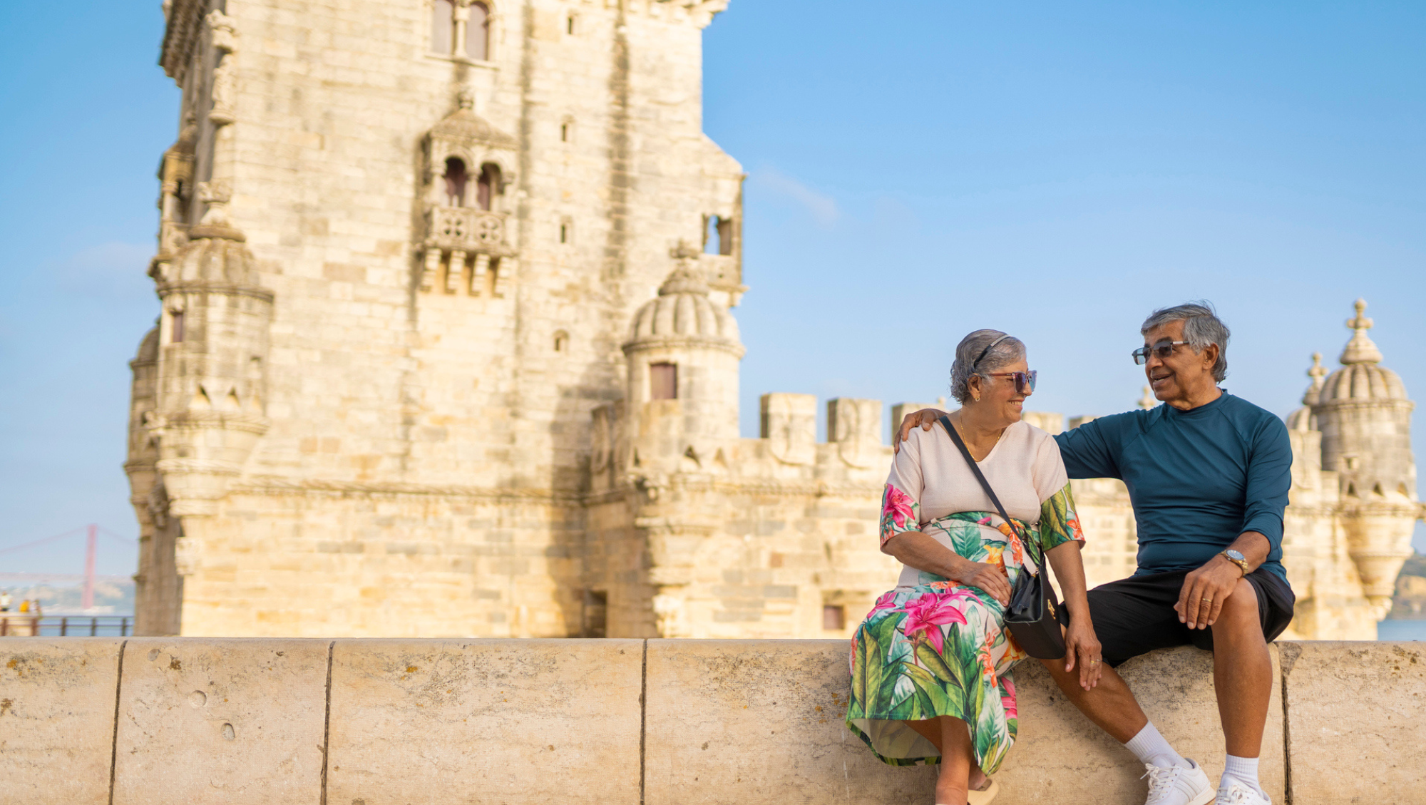 a senior couple sits in front of Belem Tower, Portugal, on vacation after asking, "Is travel insurance worth it?"