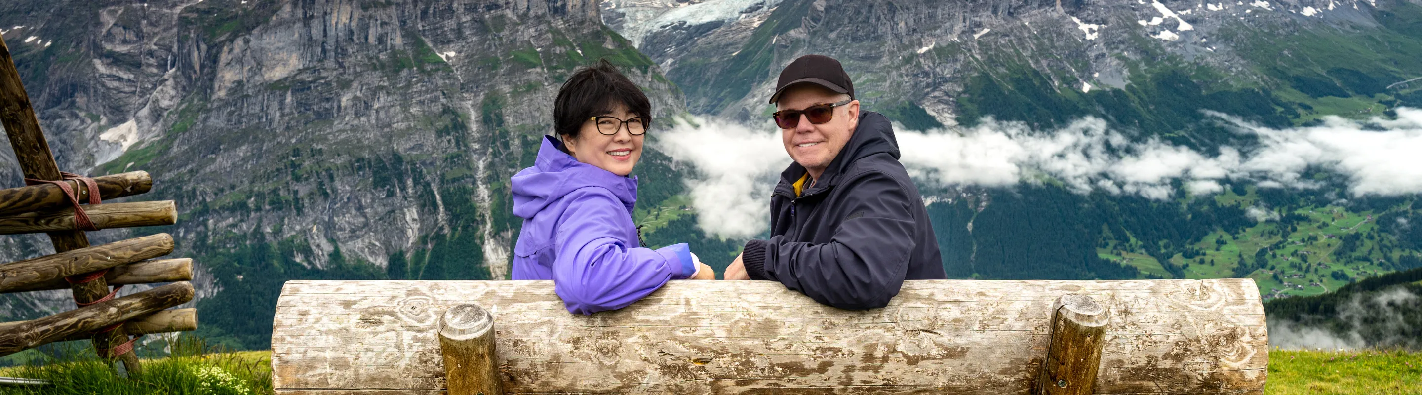 a senior, mixed-race couple sits on a wood bench with mountain views, insured with travel insurance for Switzerland 