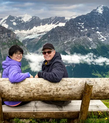 a senior, mixed-race couple sits on a wood bench with mountain views, insured with travel insurance for Switzerland 