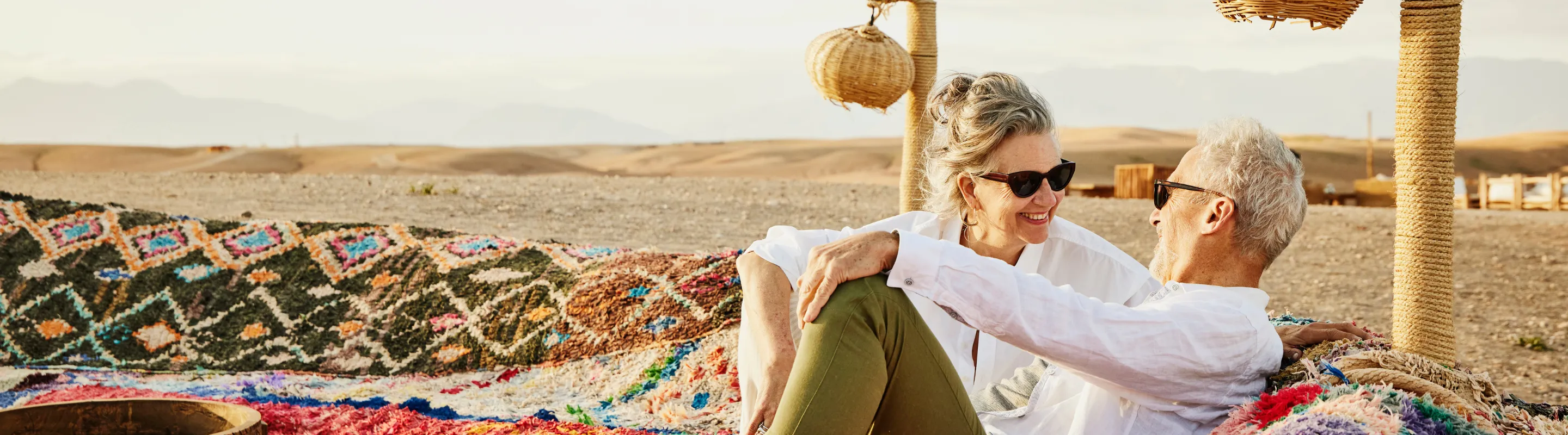 a senior couple relaxes on woven cloth in the desert, insured with travel insurance for Morocco 