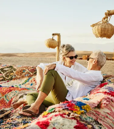 a senior couple relaxes on woven cloth in the desert, insured with travel insurance for Morocco 