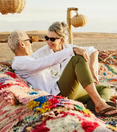 a senior couple relaxes on woven cloth in the desert, insured with travel insurance for Morocco 