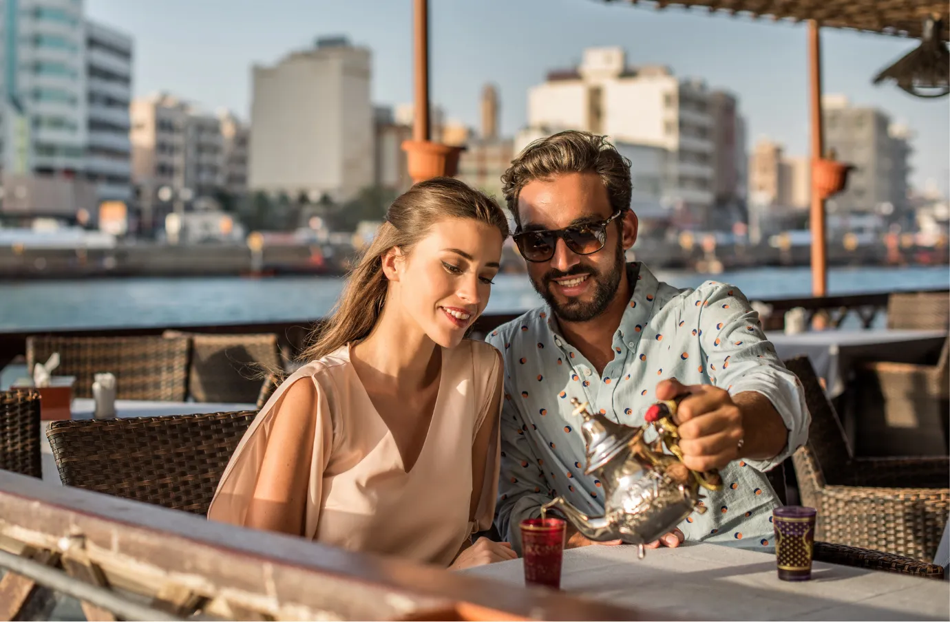 a couple with travel insurance for Dubai dine outdoors along Dubai Creek with traditional Arabic tea being poured
