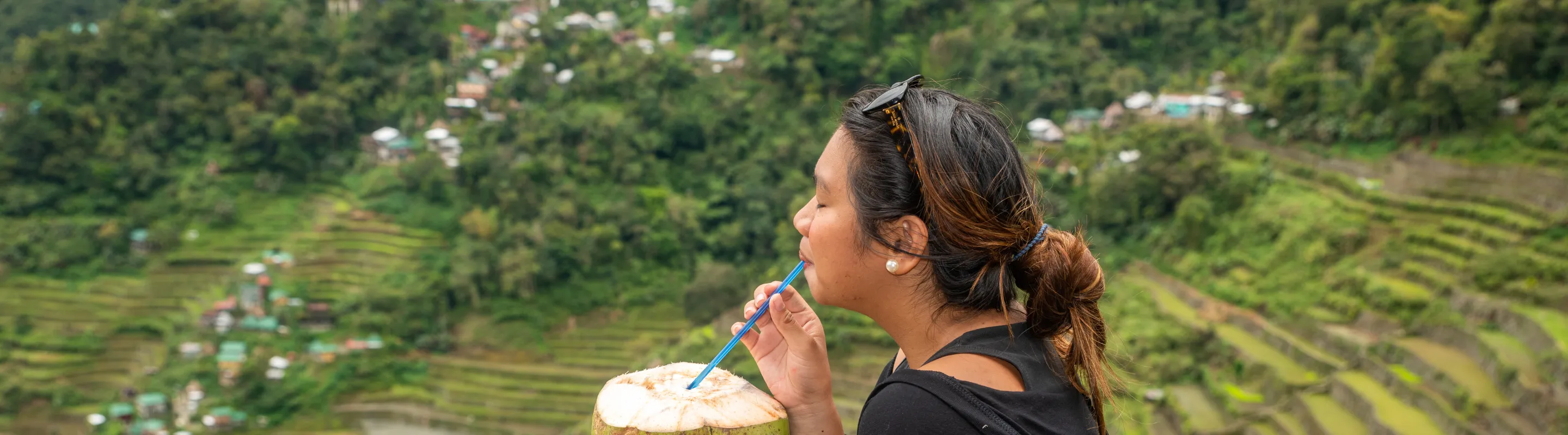 a woman with travel insurance for the Philippines drinks from coconut while overlooking a lush valley with terraced fields