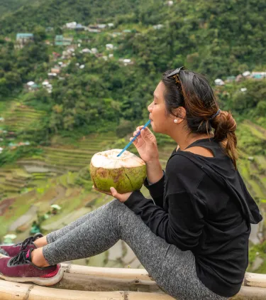a woman with travel insurance for the Philippines drinks from coconut while overlooking a lush valley with terraced fields