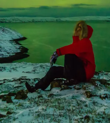 a traveler in a red jacket sits on the ground and watches the Northern Lights, insured with travel insurance for Iceland 