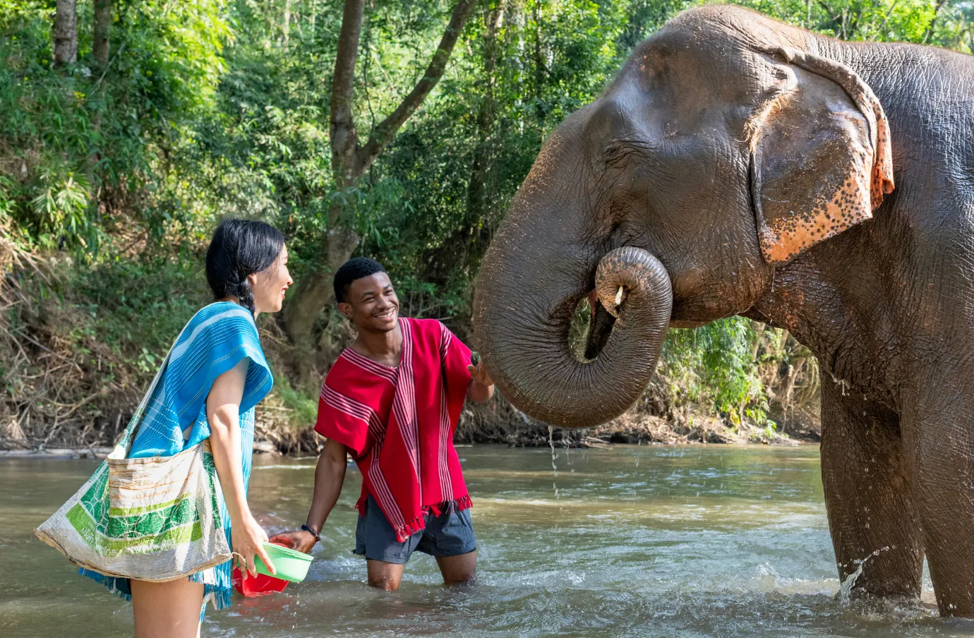 two travelers with travel insurance for Thailand bathe an elephant in a river