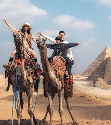 a family of three raises their arms as they ride camels at the Pyramids of Giza, insured with travel insurance for Egypt