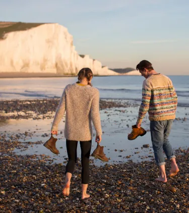 two travelers with travel insurance for the UK walk barefoot along a pebble beach beneath the Seven Sisters white cliffs