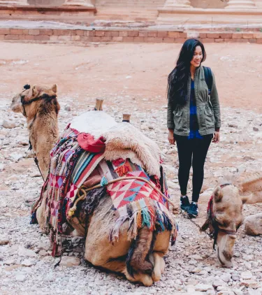 a young woman with travel insurance for Jordan cautiously stands by two camels in front of Petra