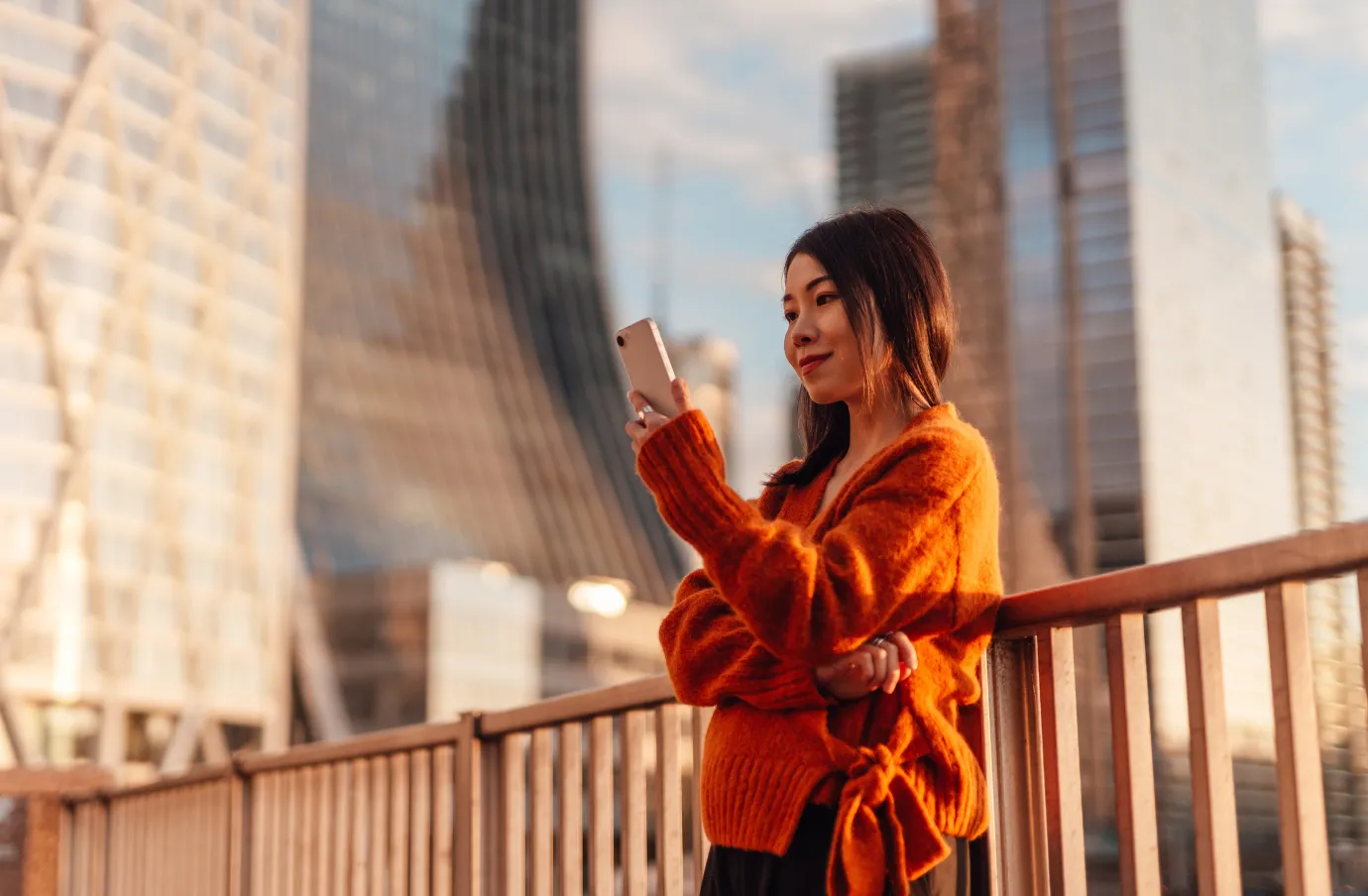 a young woman looks at her phone in a city with skyscrapers at sunset after reading our travel insurance FAQs