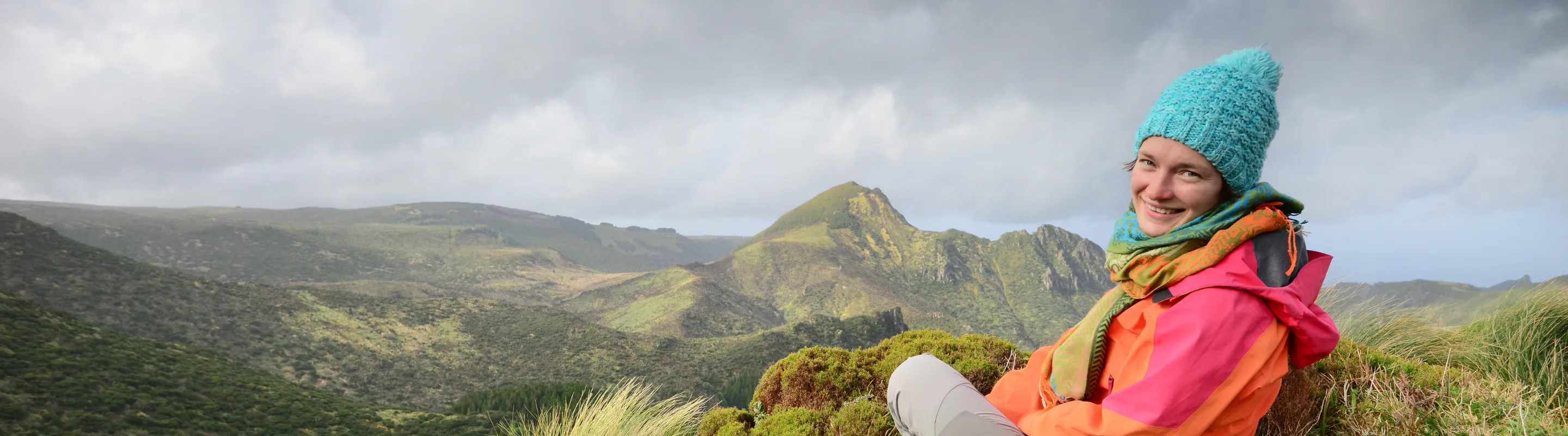 a young woman in warm clothes sits on the grass during her hike in the Azores, insured with travel insurance for Portugal