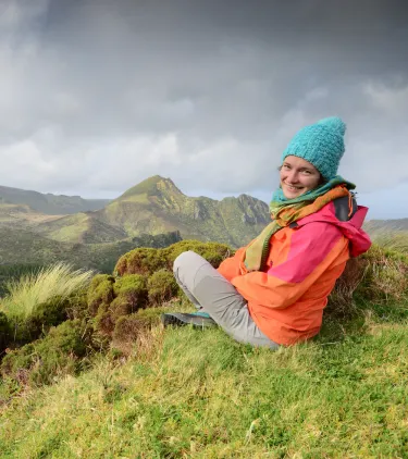 a young woman in warm clothes sits on the grass during her hike in the Azores, insured with travel insurance for Portugal
